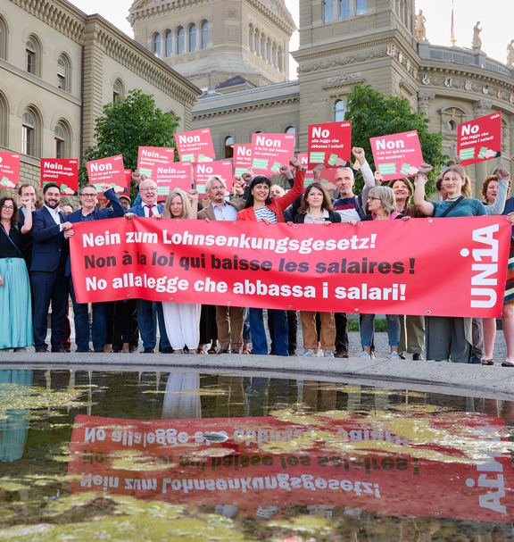 Un gruppo di persone si trova davanti al Palazzo federale con un grande striscione rosso con la scritta «No alla legge che abbassa i salari!» e cartelli in tre lingue con lo stesso messaggio; molte persone alzano il pugno in segno di protesta.