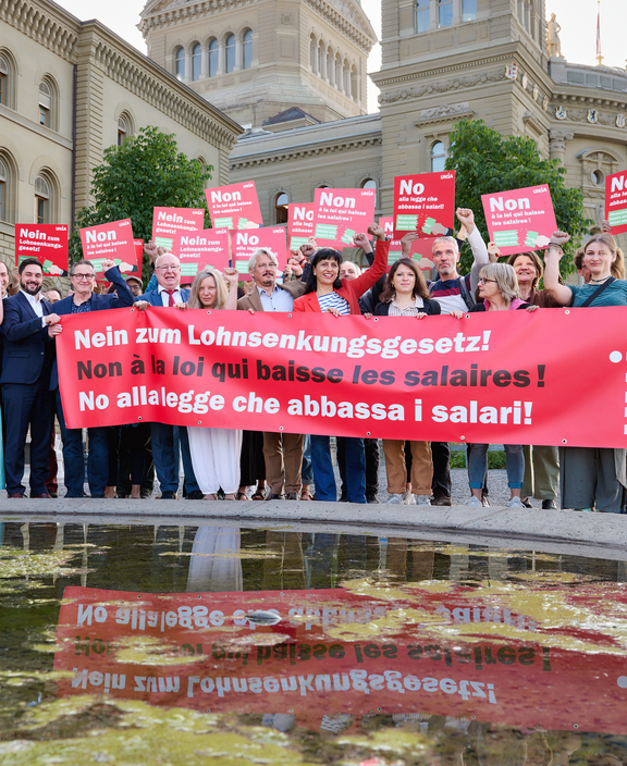 Eine Gruppe von Menschen steht vor dem Bundeshaus und hält ein grosses rotes Tranparent mit der Aufschrift «Nein zum Lohnsenkungsgesetz!» sowie Plakate in drei Sprachen mit derselben Botschaft; viele heben die Faust als Zeichen des Protests.