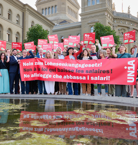 Un groupe de personnes se tient devant le Palais fédéral avec une grande banderole rouge portant l’inscription «Non à la loi qui baisse les salaires!» ainsi que des affiches en trois langues portant le même message; plusieurs lèvent le poing en signe de protestation. 