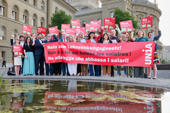 Eine Gruppe von Menschen steht vor dem Bundeshaus und hält ein grosses rotes Tranparent mit der Aufschrift «Nein zum Lohnsenkungsgesetz!» sowie Plakate in drei Sprachen mit derselben Botschaft; viele heben die Faust als Zeichen des Protests.