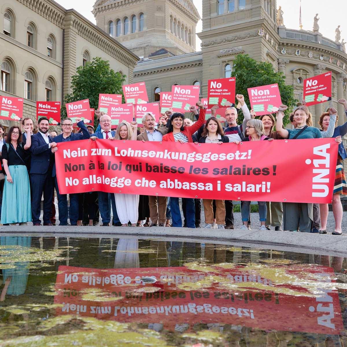 Eine Gruppe von Menschen steht vor dem Bundeshaus und hält ein grosses rotes Tranparent mit der Aufschrift «Nein zum Lohnsenkungsgesetz!» sowie Plakate in drei Sprachen mit derselben Botschaft; viele heben die Faust als Zeichen des Protests.