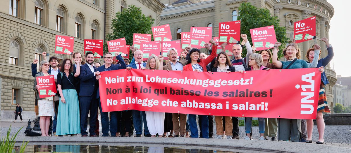 Eine Gruppe von Menschen steht vor dem Bundeshaus und hält ein grosses rotes Tranparent mit der Aufschrift «Nein zum Lohnsenkungsgesetz!» sowie Plakate in drei Sprachen mit derselben Botschaft; viele heben die Faust als Zeichen des Protests.