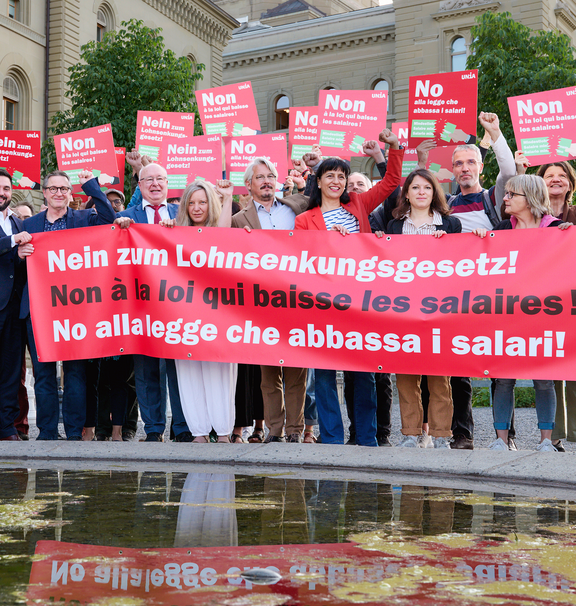 Eine Gruppe von Menschen steht vor dem Bundeshaus und hält ein grosses rotes Tranparent mit der Aufschrift «Nein zum Lohnsenkungsgesetz!» sowie Plakate in drei Sprachen mit derselben Botschaft; viele heben die Faust als Zeichen des Protests.