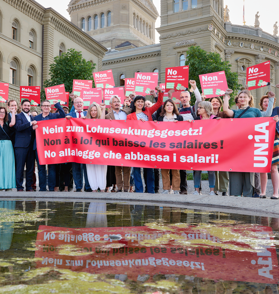 Un gruppo di persone si trova davanti al Palazzo federale con un grande striscione rosso con la scritta «No alla legge che abbassa i salari!» e cartelli in tre lingue con lo stesso messaggio; molte persone alzano il pugno in segno di protesta.