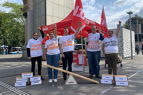 5 Menschen in bemalten T-Shirts von dem Luzerner Bahnhof vor einer symbolischen Waage: Teuerung, Krankenkasse, Miete etc auf der einen Seite; Lohn auf der anderen: 