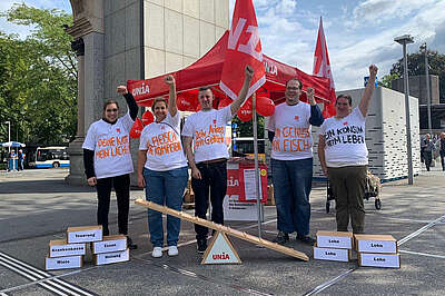 5 Menschen in bemalten T-Shirts von dem Luzerner Bahnhof vor einer symbolischen Waage: Teuerung, Krankenkasse, Miete etc auf der einen Seite; Lohn auf der anderen: 
