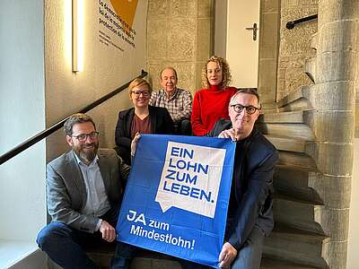 5 membri del comitato d'iniziativa siedono su una scala e tengono nella telecamera una bandiera con la scritta “Un salario per vivere” – motivo e slogan della campagna.