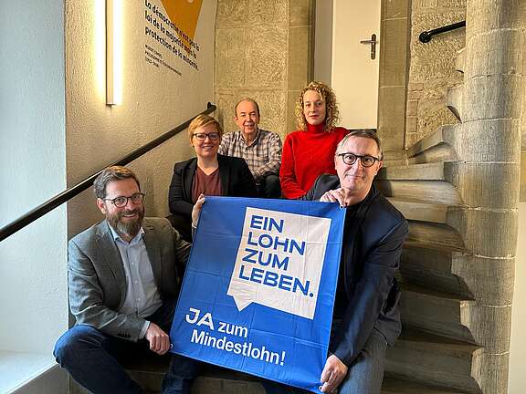 5 membri del comitato d'iniziativa siedono su una scala e tengono nella telecamera una bandiera con la scritta “Un salario per vivere” – motivo e slogan della campagna.