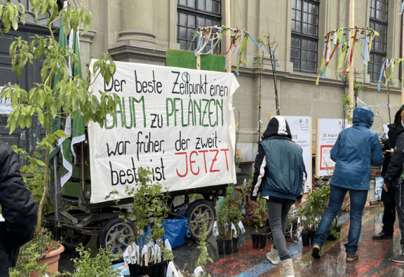 Mehrere Personen stehen bei Regen vor einem mit Pflanzen geschmückten Transparent mit dem Text: «Der beste Zeitpunkt einen Baum zu pflanzen war früher, der zweitbeste ist JETZT»