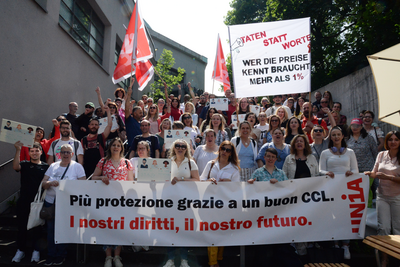 150 persone su una scala innalzano cartelli e bandiere. La prima fila regge uno striscione con la scritta: «Più protezione grazie a un CCL forte: il nostro futuro, i nostri diritti».