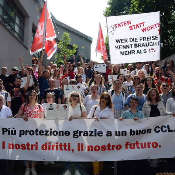 Un gruppo numeroso di persone riunite all'aperto, sorridenti e con le braccia alzate, partecipa a una manifestazione sindacale. In primo piano, uno striscione bianco con la scritta in italiano e tedesco: 'Più protezione grazie a un buon CCL. I nostri diritti, il nostro futuro.' e 'WER DIE PREISE KENNT BRAUCHT MEHR ALS 1%'. Sono presenti anche bandiere rosse con il logo del sindacato Unia.