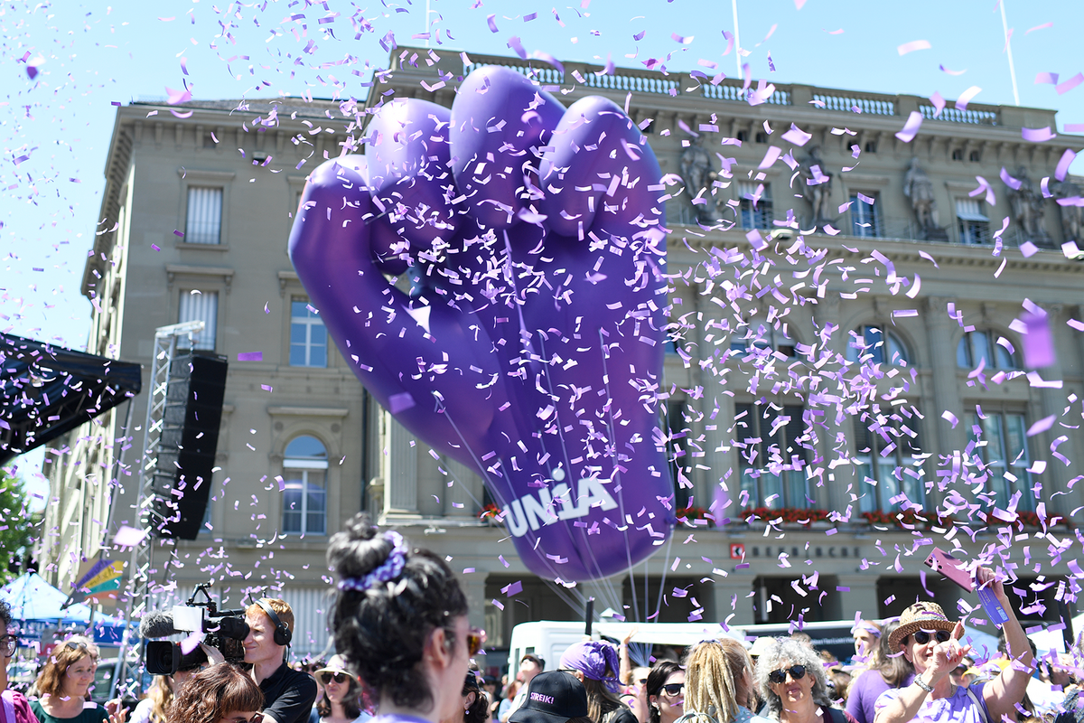 Un gros poing fermé violet gonflable avec le logo d'Unia s'élève dans des confettis au-dessus d'une manifestation.