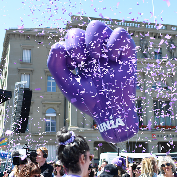 Un énorme poing gonflable violet marqué du logo d'Unia s'élève au-dessus d'une manifestation féministe