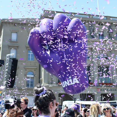Un gros poing fermé violet gonflable avec le logo d'Unia s'élève dans des confettis au-dessus d'une manifestation.