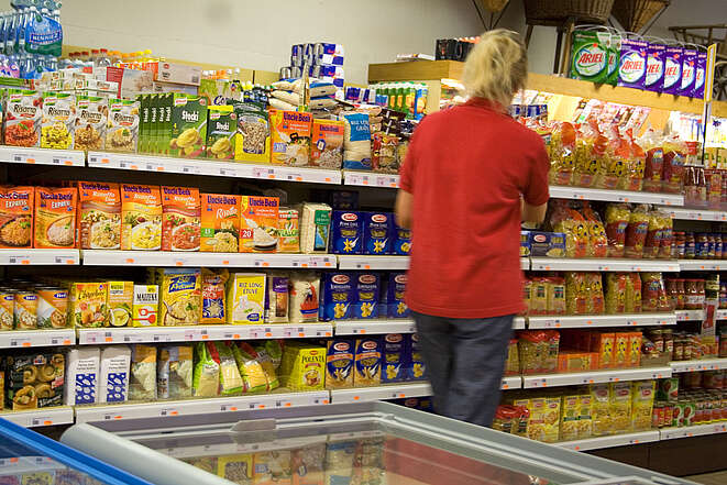 Eine Supermarkt-Angestellte in rotem Uniform-Shirt steht vor einem Regal, das mit verschiedenen Nudel-, Reis- und Instantgerichten gefüllt ist. 