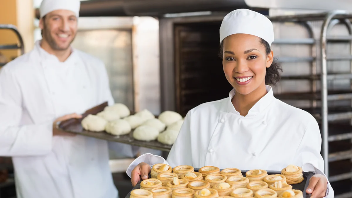 Un panettiere e una panettiera in abiti da lavoro bianchi presentano preparazioni fresche e dolci cotti in un forno. 