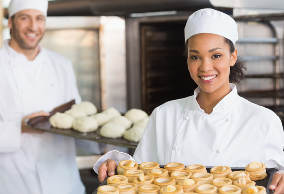 Un panettiere e una panettiera in abiti da lavoro bianchi presentano preparazioni fresche e dolci cotti in un forno. 