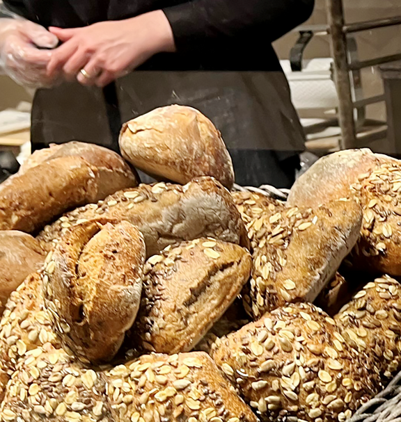 Auslage in einer Bäckerei mit verschiedenen Brotsorten und Gebäck in Weidenkörben; im Hintergrund sind Hände beim Verpacken von Backwaren zu sehen.