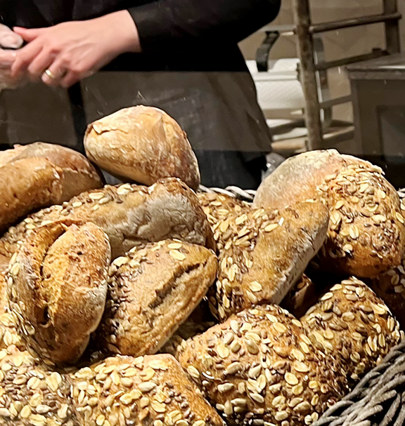 Esposizione in una panetteria con diverse varietà di pane e prodotti da forno disposti in cesti di vimini; sullo sfondo si vedono mani che confezionano prodotti da forno.