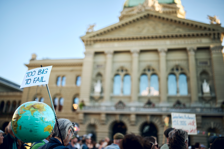 Une personne porte sur l’épaule une perche avec un globe terrestre gonflable et une pancarte portant l’inscription «Too big to fail». À l’arrière-plan, le Palais fédéral à Berne et d’autres manifestant-e-s.