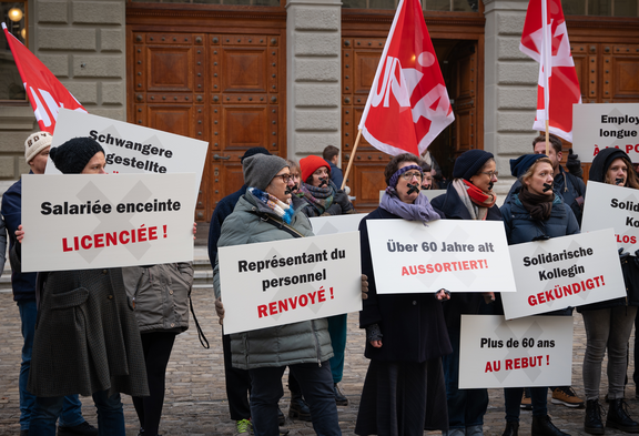 Des manifestant-e-s brandissent des pancartes avec des revendications contre les licenciements, notamment pour les femmes enceintes et les salarié-e-s âgés. Des drapeaux Unia sont visibles à l'arrière-plan. 