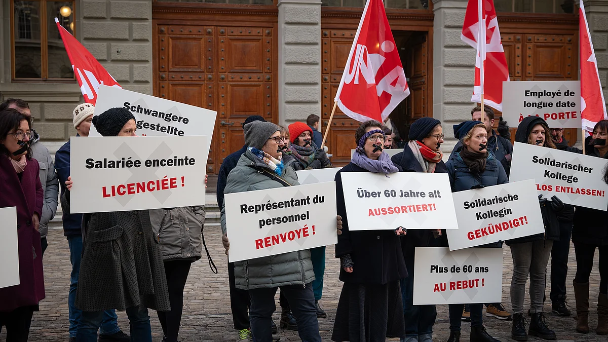 Un gruppo di manifestanti mostra cartelli con rivendicazioni contro i licenziamenti, soprattutto a tutela delle donne incinte e del personale più anziano. Sullo sfondo sventolano le bandiere di Unia.