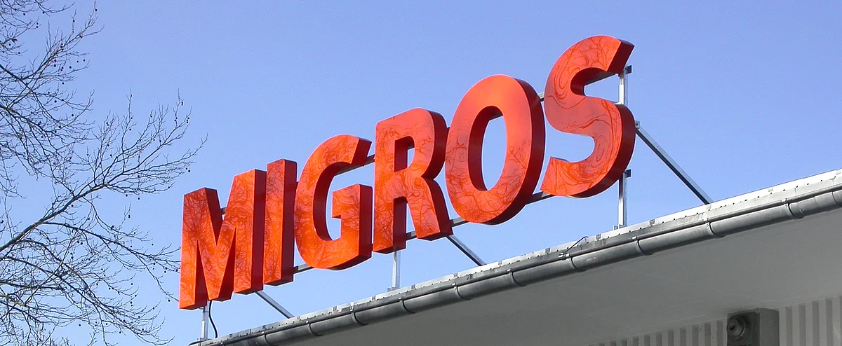 De grandes lettres orange « Migros » sur le toit d’un magasin. 