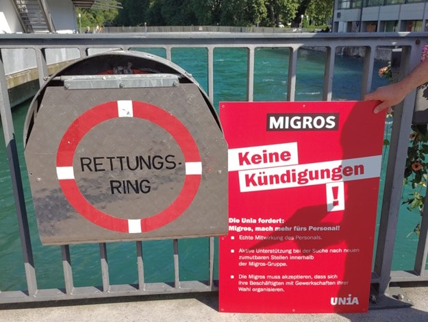 Ein Protestschild, auf dem die Forderungen der Unia an die Migrosleitung aufgelistet ist, steht neben einem Rettungsring-Kasten.