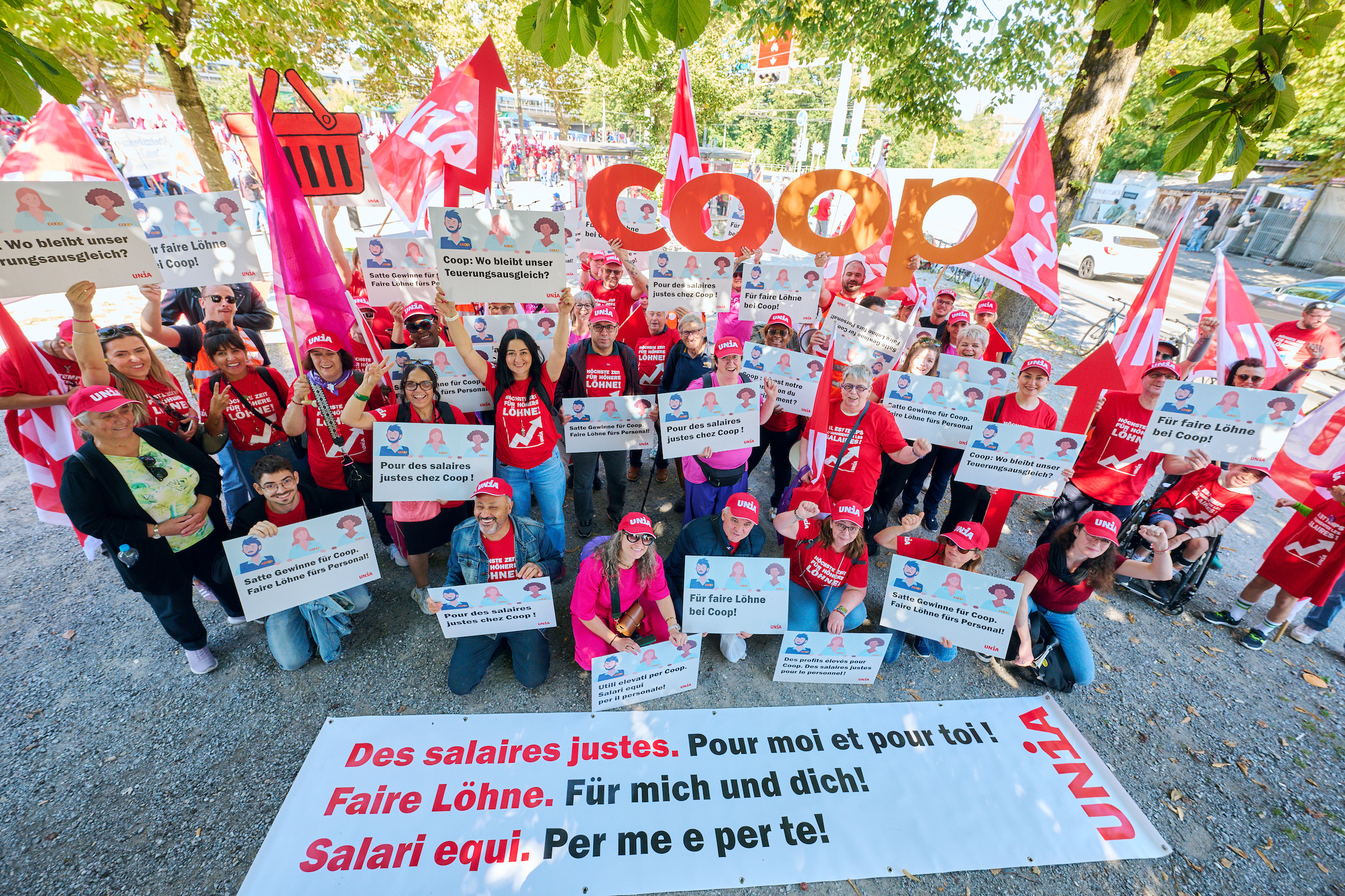 Un groupe d’employé-e-s Coop brandissent des drapeaux, le logo Coop et des pancartes avec leurs revendications aux employeurs: «Des bénéfices énormes pour Coop. Des salaires équitables chez Coop». Sur une banderole posée au sol, on peut lire dans trois langues «Des salaires équitables. Pour moi et pour toi!».