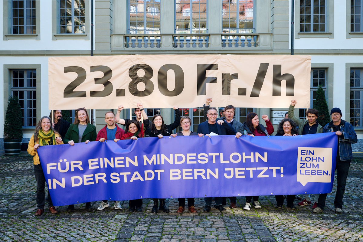 Im Rahmen der Einreichung einer Initiative für einen Mindestlohn in der Stadt Bern steht eine Gruppe von etwa 15 Personen aus dem Bündnis für diese Initiative vor einem historischen Gebäude in Bern und hält zwei Banner hoch. Das obere Banner zeigt in grossen schwarzen Buchstaben die Forderung "23.80 Fr./h". Das untere Banner ist violett und trägt die deutsche Aufschrift "FÜR EINEN MINDESTLOHN IN DER STADT BERN JETZT!" Auf der rechten Seite des Banners befindet sich ein Emblem mit dem deutschen Text "EIN LOHN ZUM LEBEN". Einige Personen in der Gruppe heben die Faust und lächeln. 