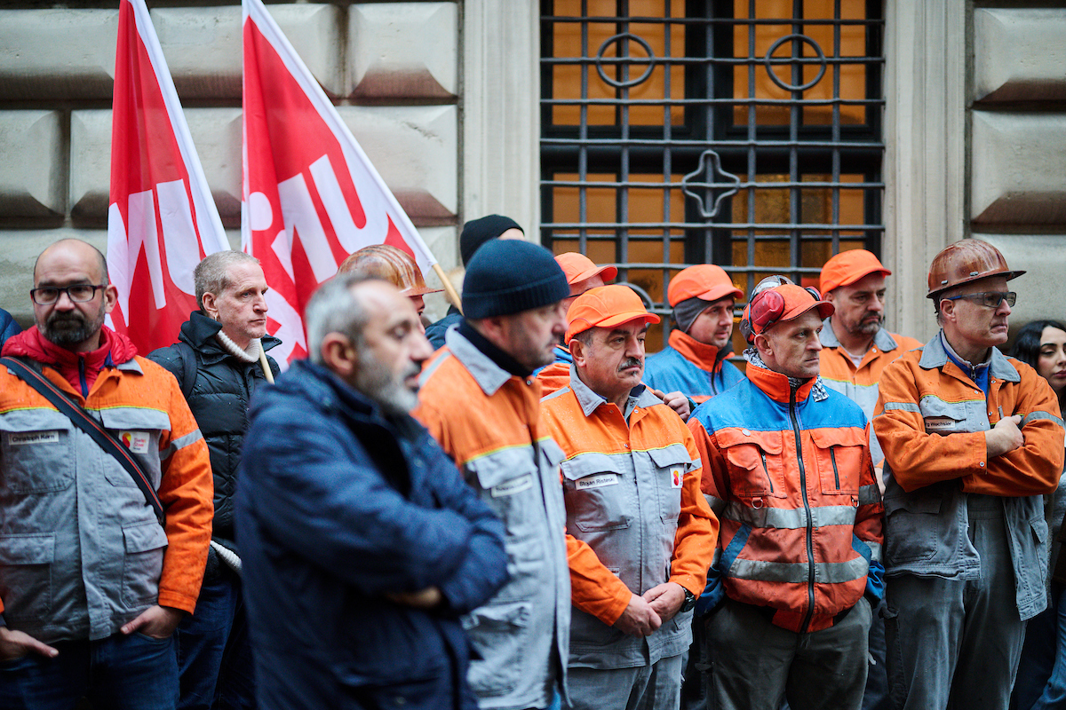 Beschäftigten von Steeltec in Arbeitskleidung und Warnwesten, einige mit Helmen, stehen eng beisammen vor dem Luzerner Parlamentsgebäude mit ernsten und besorgten Gesichtern. Im Hintergrund sind Unia-Fahnen sichtbar.