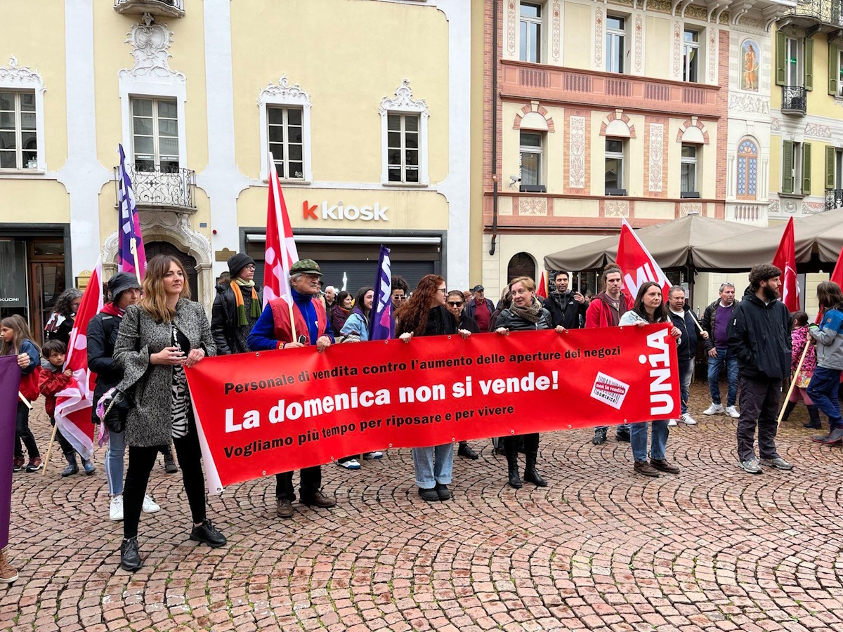 Persone partecipano a una manifestazione in una piazza con pavimentazione in pietra (sampietrini). Sorreggono uno striscione rosso con la scritta: «La domenica non si vende! Vogliamo più tempo per riposare e per vivere.» Sullo sfondo si vedono edifici storici con facciate decorate e un «kiosk». I partecipanti sventolano bandiere rosse e viola."