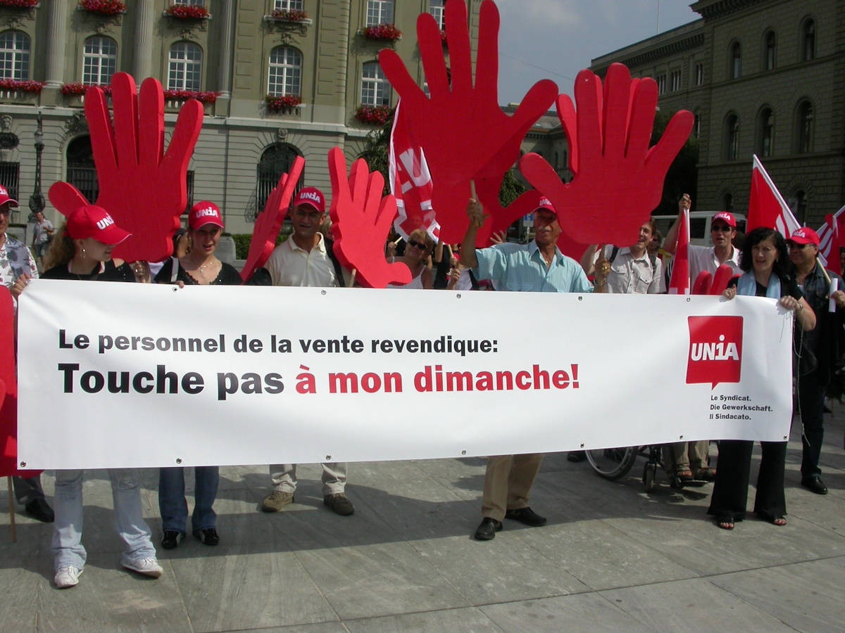 Un groupe de personnes manifeste avec une grande banderole et de fausses mains rouges contre l’élargissement des horaires d’ouverture dominicaux. Le slogan suivant y figure: «Le personnel de vente revendique: touche pas à mon dimanche!» Une partie des manifestant-e-s portent des casquettes du syndicat. On aperçoit des bâtiments à l’arrière-plan.