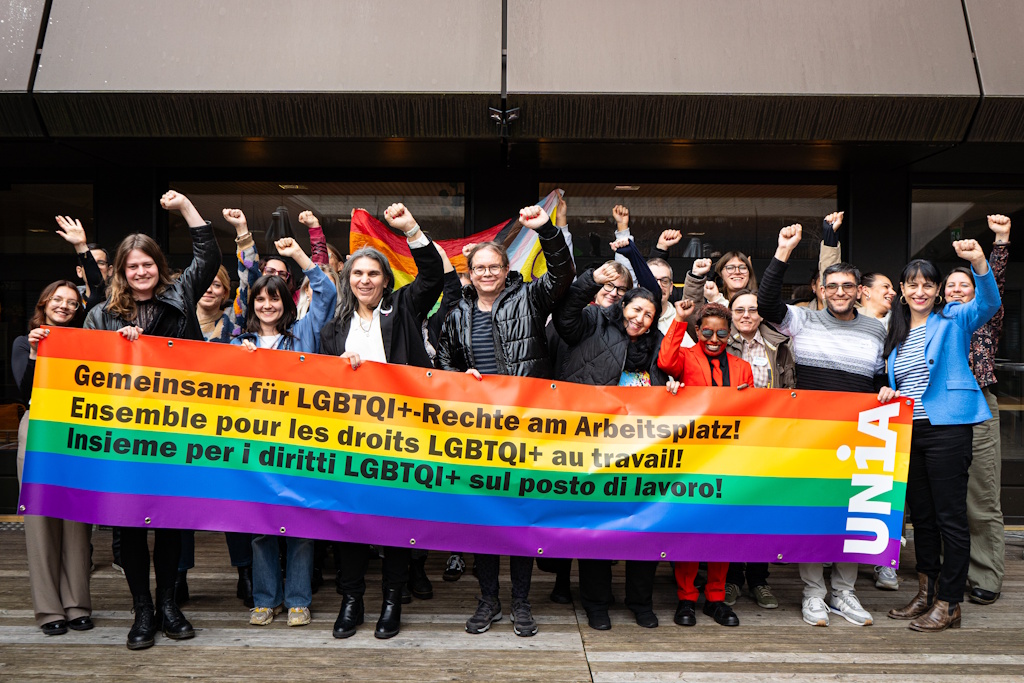 Un groupe de personnes tient une bannière arc-en-ciel avec l'inscription «Ensemble pour les droits LGBTQI+ au travail!» et lève le poing avec détermination.