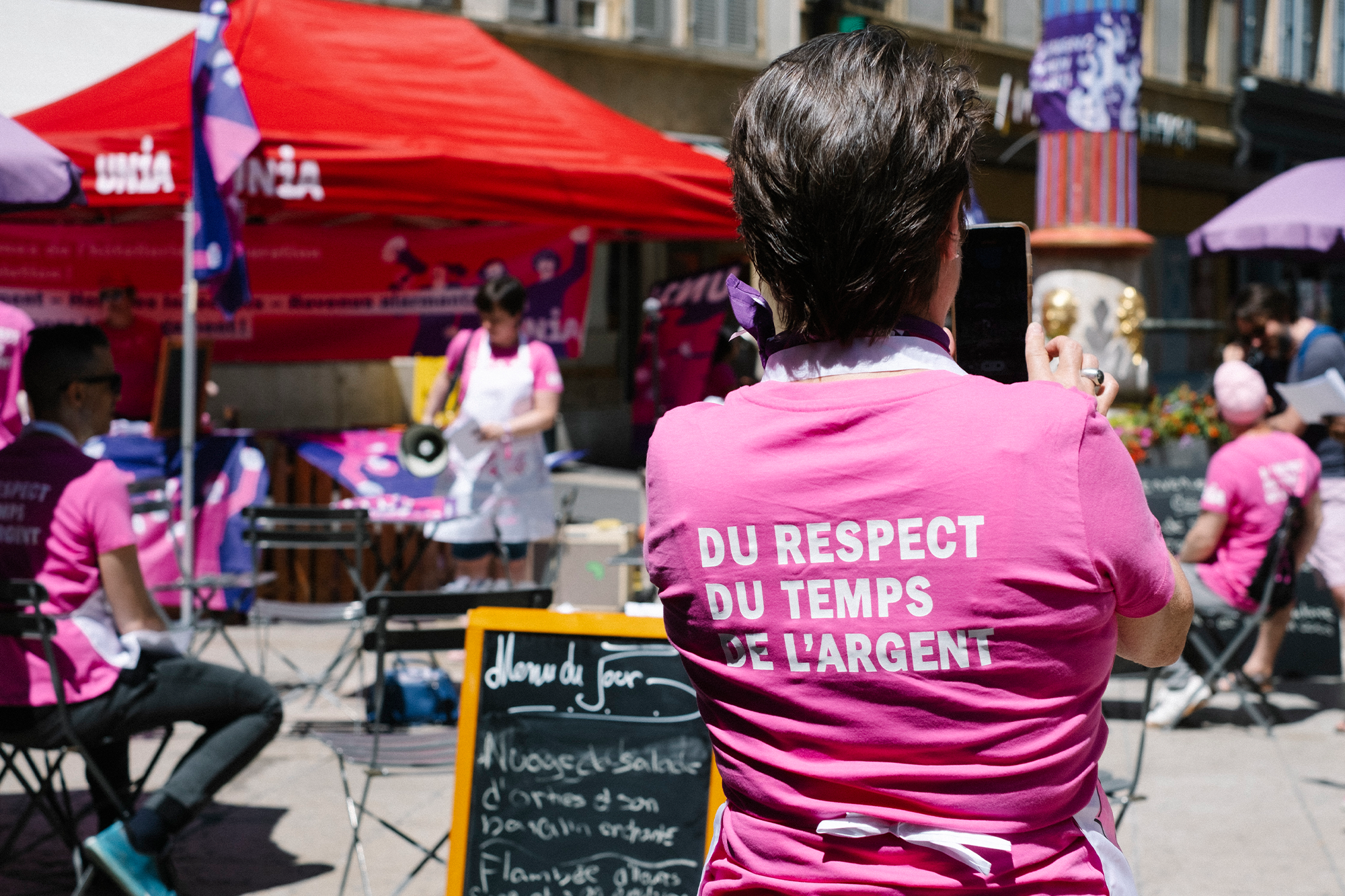 La photo représente une manifestation avec des personnes vêtues de t-shirts roses et arborant des symboles féministes. Le T-shirt de la personne au premier plan porte l’inscription « Du respect, du temps, de l’argent ! ». On distingue au fond une terrasse de restaurant. L’esprit qui transparaît de la photo est déterminé et combatif.