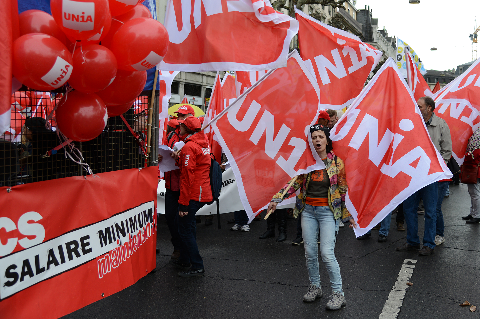 Il s’agit d’une action de rue avec des militant-e-s Unia habillés en rouge et avec des drapeaux Unia. On peut lire sur une banderole «Salaire minimum maintenant».