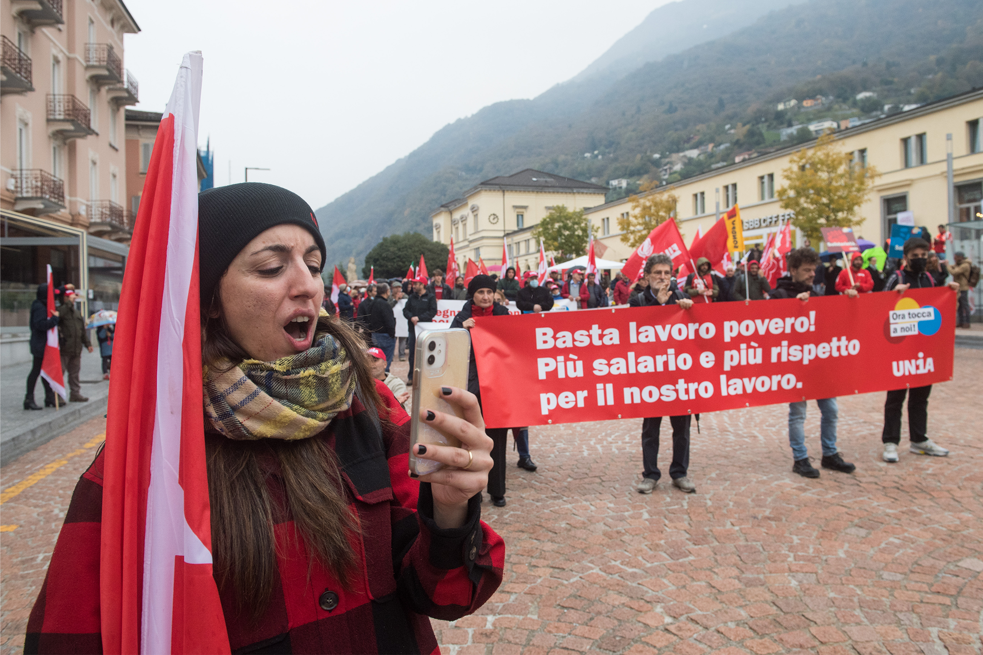 La foto mostra un'azione di strada con attivisti di Unia vestiti di rosso e con bandiere di Unia. Uno striscione recitava “Basta lavoro povero! Più salario e più rispetto per il nostro lavoro”.