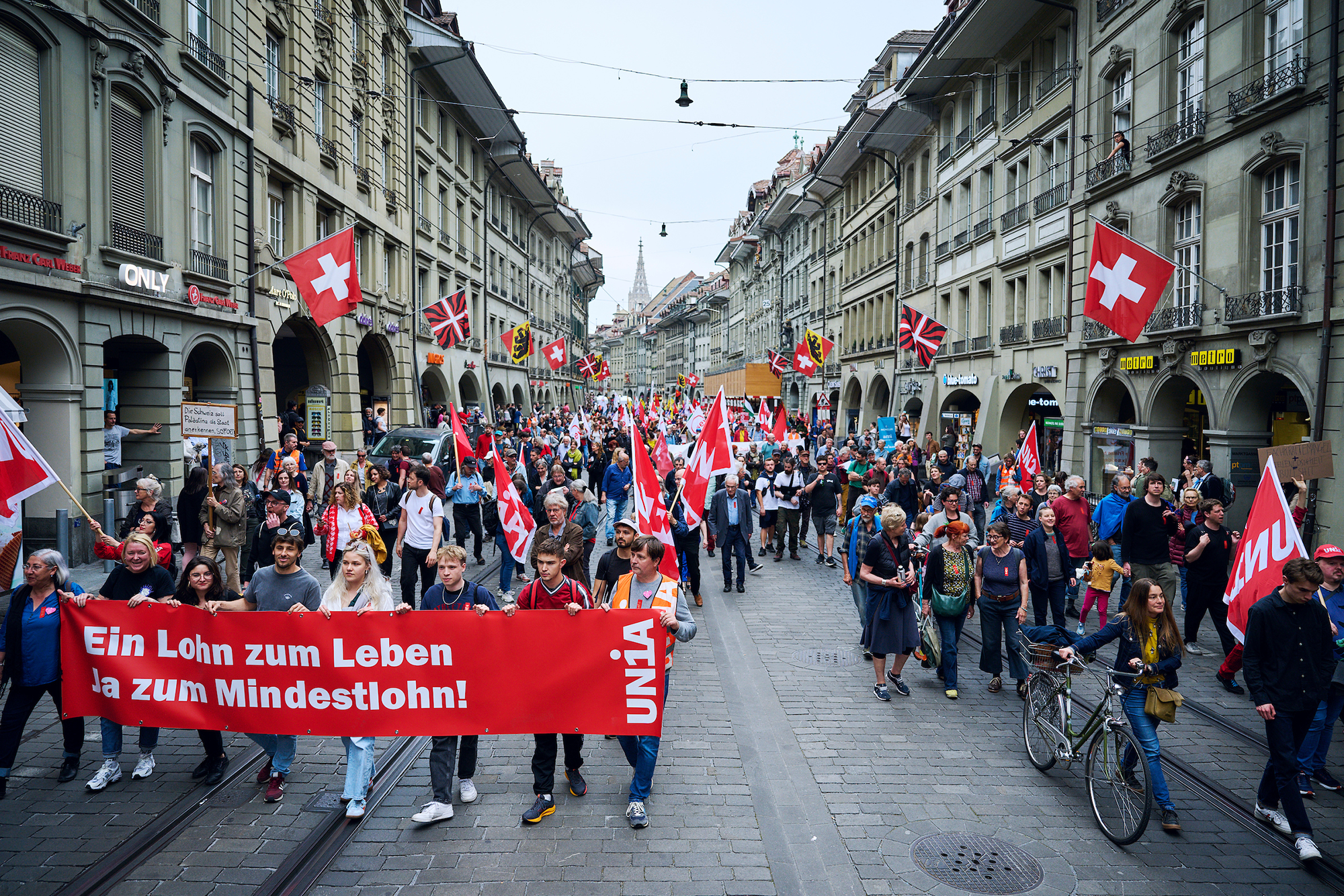 Es handelt sich um eine Strassenaktion mit rot gekleideten Unia-Aktivistinnen mit Unia-Fahnen. Auf einem Transparent ist zu lesen: "Ein Lohn zum Leben. Ja zum Mindestlohn!".