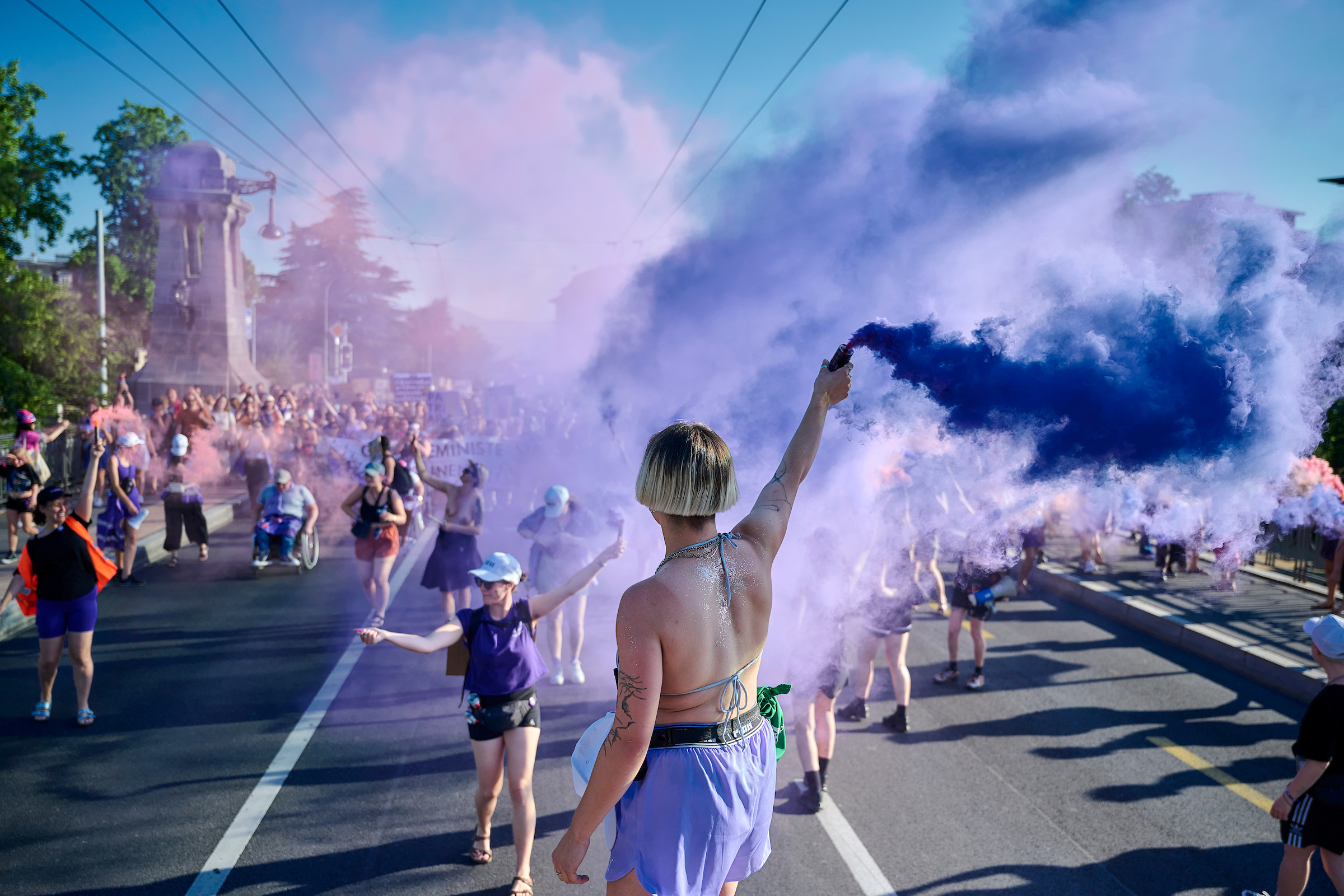 Scène animée sur le Pont Chaudron à Lausanne dans le cadre d'une manifestation de la grève des femmes. Au premier plan, une personne aux cheveux courts et blonds agite une bombe fumigène couleur lila. De la fumée violette enveloppe l'environnement, tandis que la foule, y compris une personne en fauteuil roulant, défile.