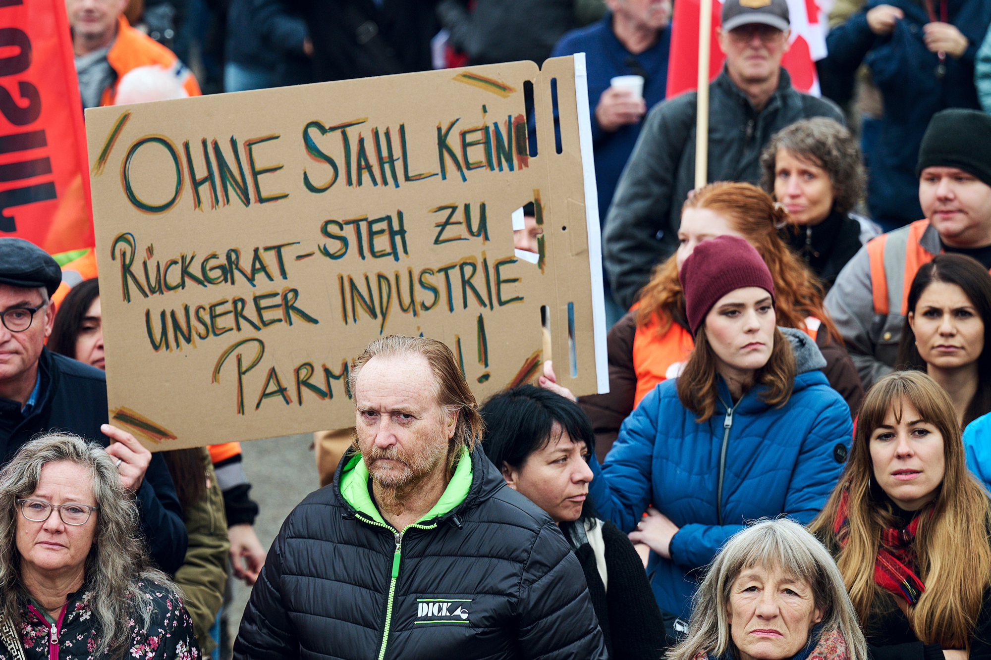 Des personnes lors d'une manifestation ; au milieu, un panneau en carton écrit à la main avec les mots: «Sans acier, pas de colonne vertébrale. Tiens-toi à notre industrie, Parmelin».