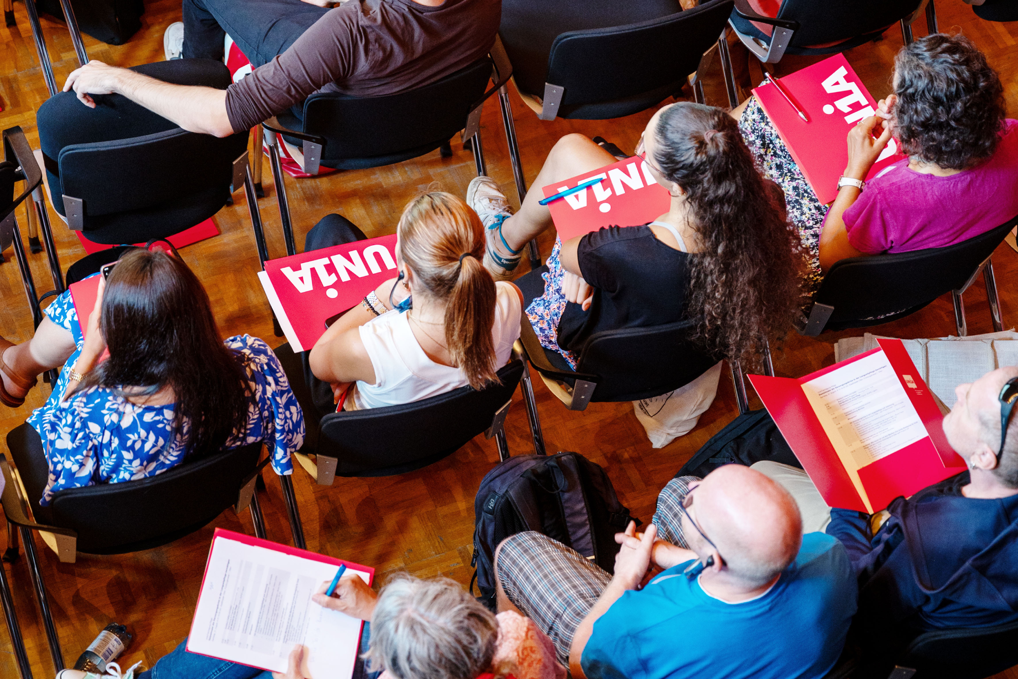 Menschen sitzen in einem Theatersaal, halten rote Mappen mit Unia darauf. Einige schreiben oder lesen Unterlagen. Sicht von oben.