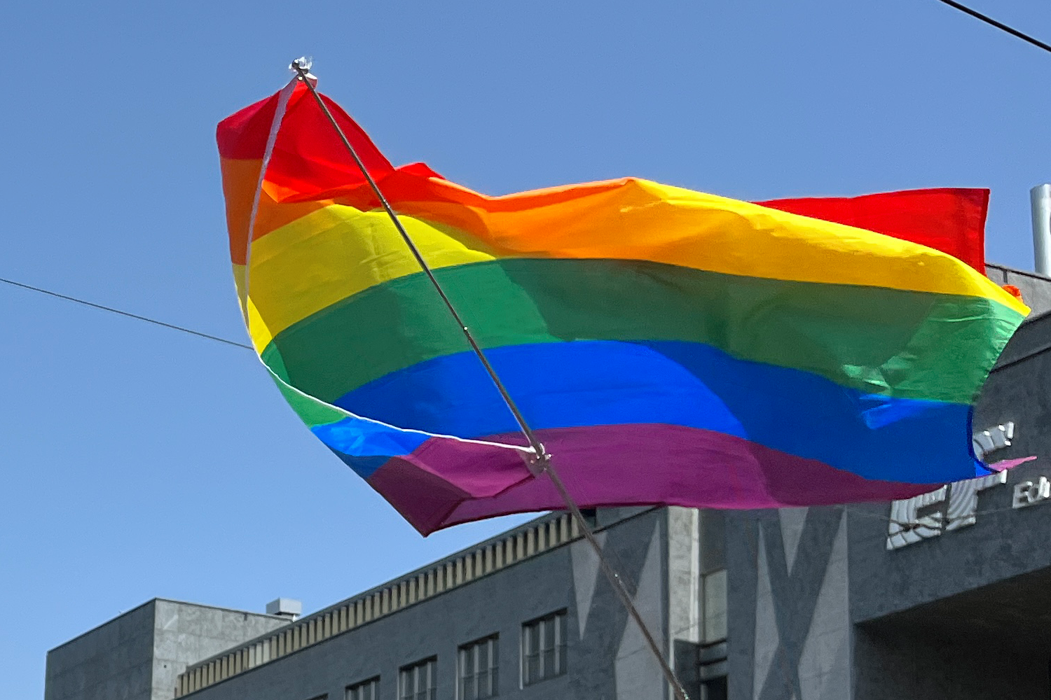 Eine Regenbogenflagge weht vor blauem Himmel, mit einem Gebäude im Hintergrund.