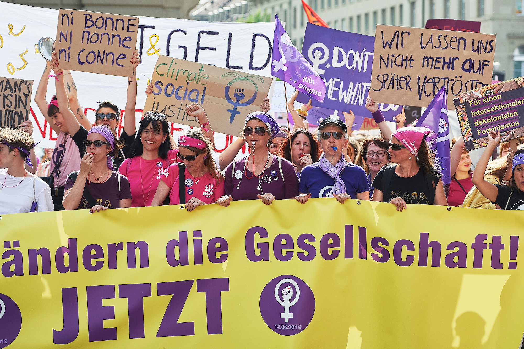 Eine feministische Demonstration mit Frauen in violett und pink, die Transparente und Schilder mit Forderungen nach Gleichberechtigung und sozialer Gerechtigkeit halten.