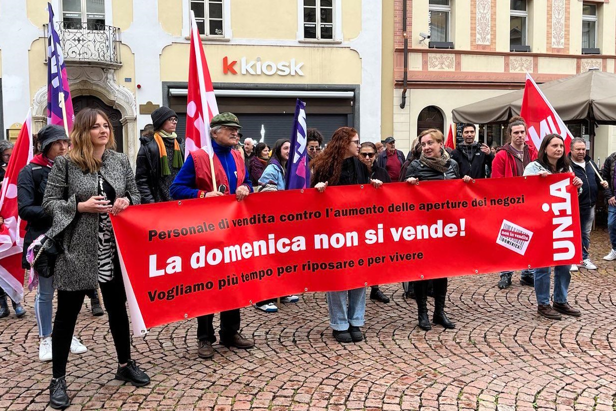 Un gruppo di manifestanti di diverse età tiene in piazza uno striscione rosso con scritta bianca e nera durante una manifestazione a Bellinzona, Svizzera