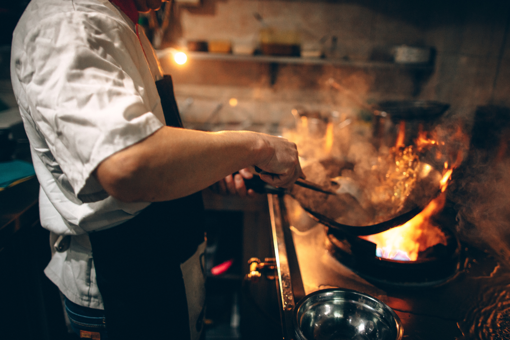 Uno chef con camicia bianca salta del cibo in una padella su una fiamma viva. Vapore e fiamme creano un’atmosfera dinamica.