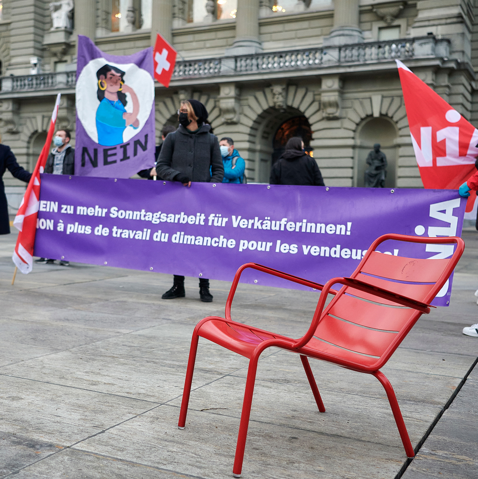 Eine Gruppe von Unia-Gewerkschafter:innen nimmt an einer Kundgebung vor dem Bundeshaus in Bern gegen zusätzliche Sonntags- und Wochenendarbeit teil. Eine Person steht hinter einem Transparent:„Nein zu mehr Sonntagsarbeit für Verkäuferinnen!“