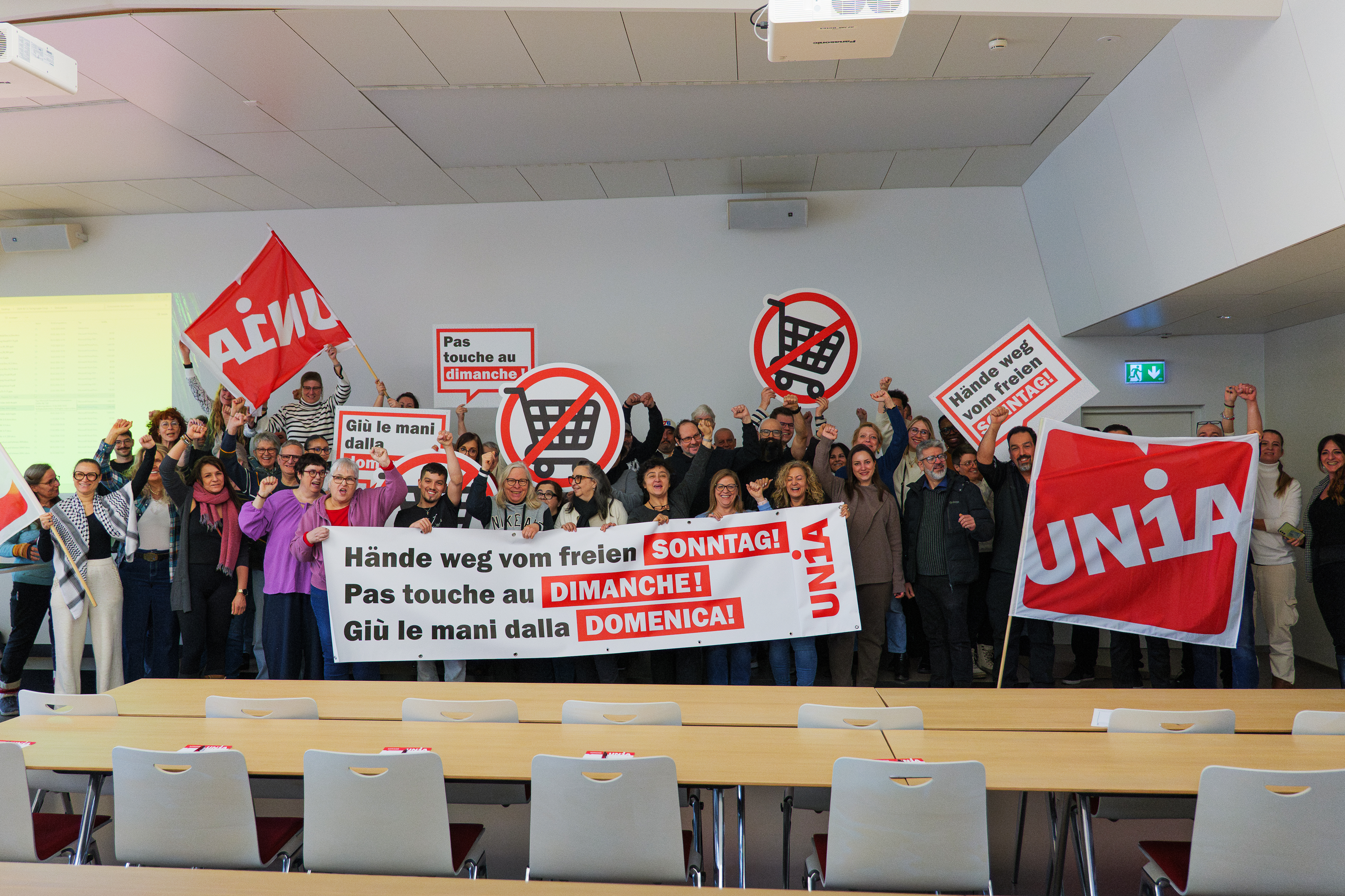 Eine Gruppe von Menschen versammelt sich hinter einem Transparent, auf dem steht «Hände weg vom freien Sonntag». Sie halten Schilder und schwenken Unia-Fahnen.